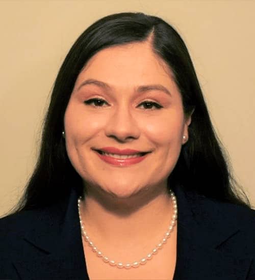 A portrait of Amy Gonzalez. She has parted, long black hair and is wearing a dark blazer against a neutral beige background.