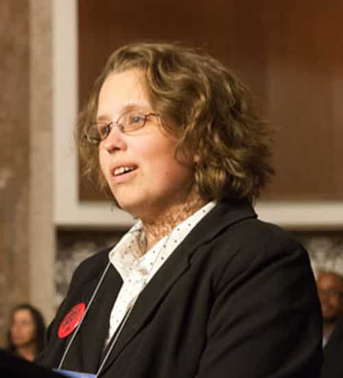 A portrait of Nicole LeBlanc. She has wavy medium-length hair, is formally dressed and appears to be addressing a meeting room of people.