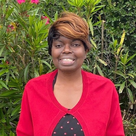 A portrait of Angela Greene. She is a black woman smiling with a colorful swoop of bangs. She is wearing a red cardigan standing in front of a clump of green hedges.